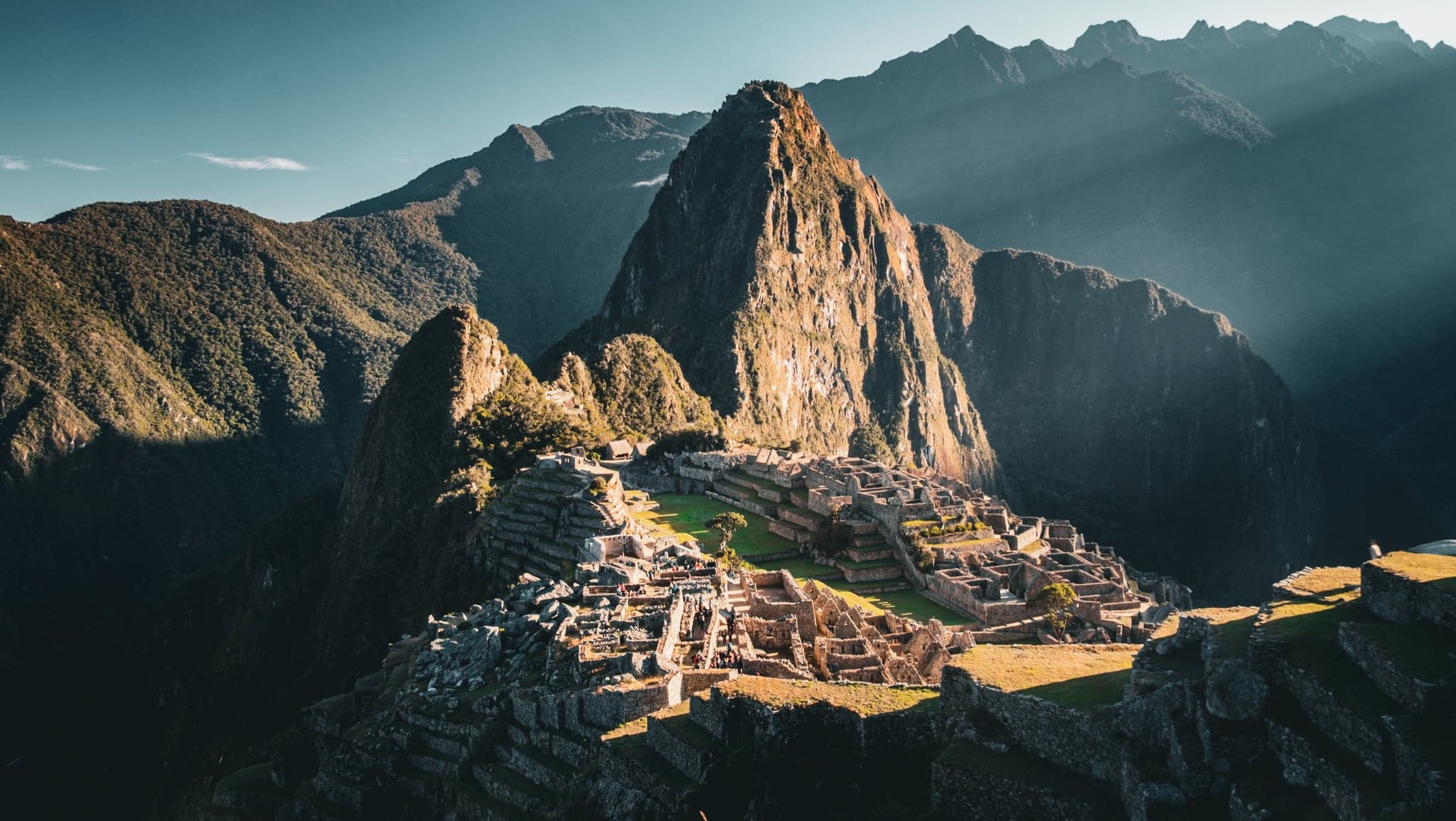 Sunlight bathes ancient stone terraces, casting long shadows across the verdant landscape, while a towering peak looms majestically in the background.