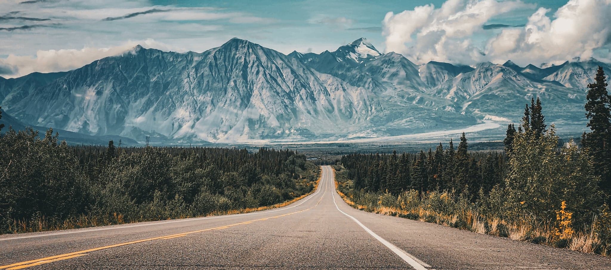 A winding road leads towards majestic mountains, their rugged peaks framed by a canopy of dramatic clouds.
