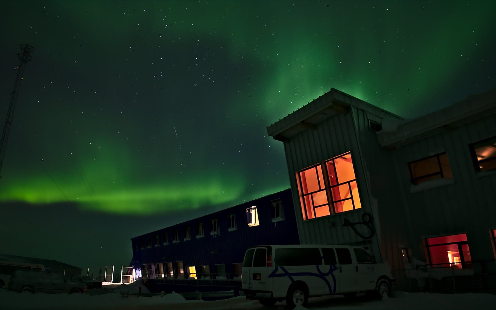 Green auroras dance above a darkened building, its windows glowing warmly, creating a stark contrast against the cold night sky.