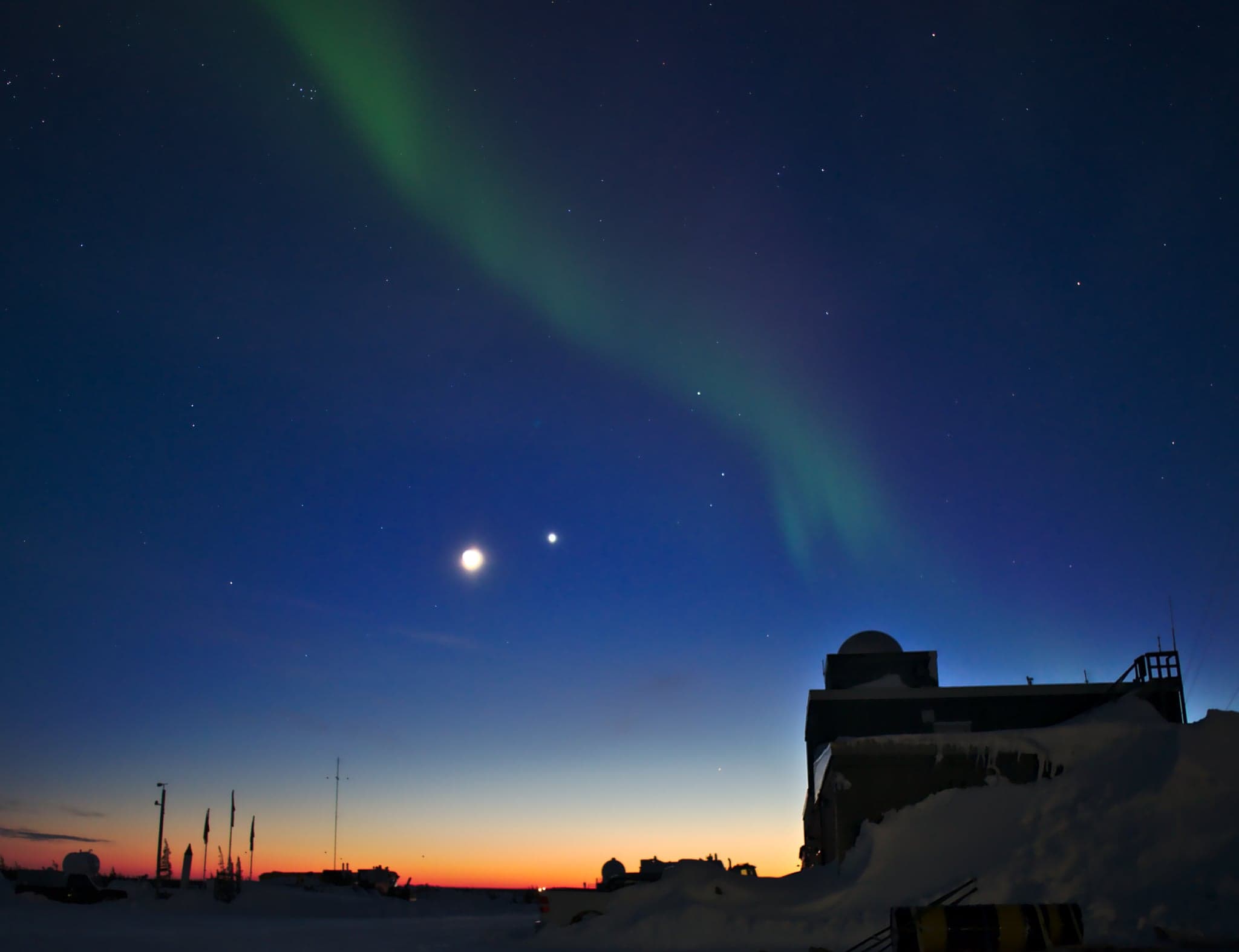 Aurora dances across a twilight sky, the crescent moon and a bright star adding celestial harmony above a snow-covered outpost.