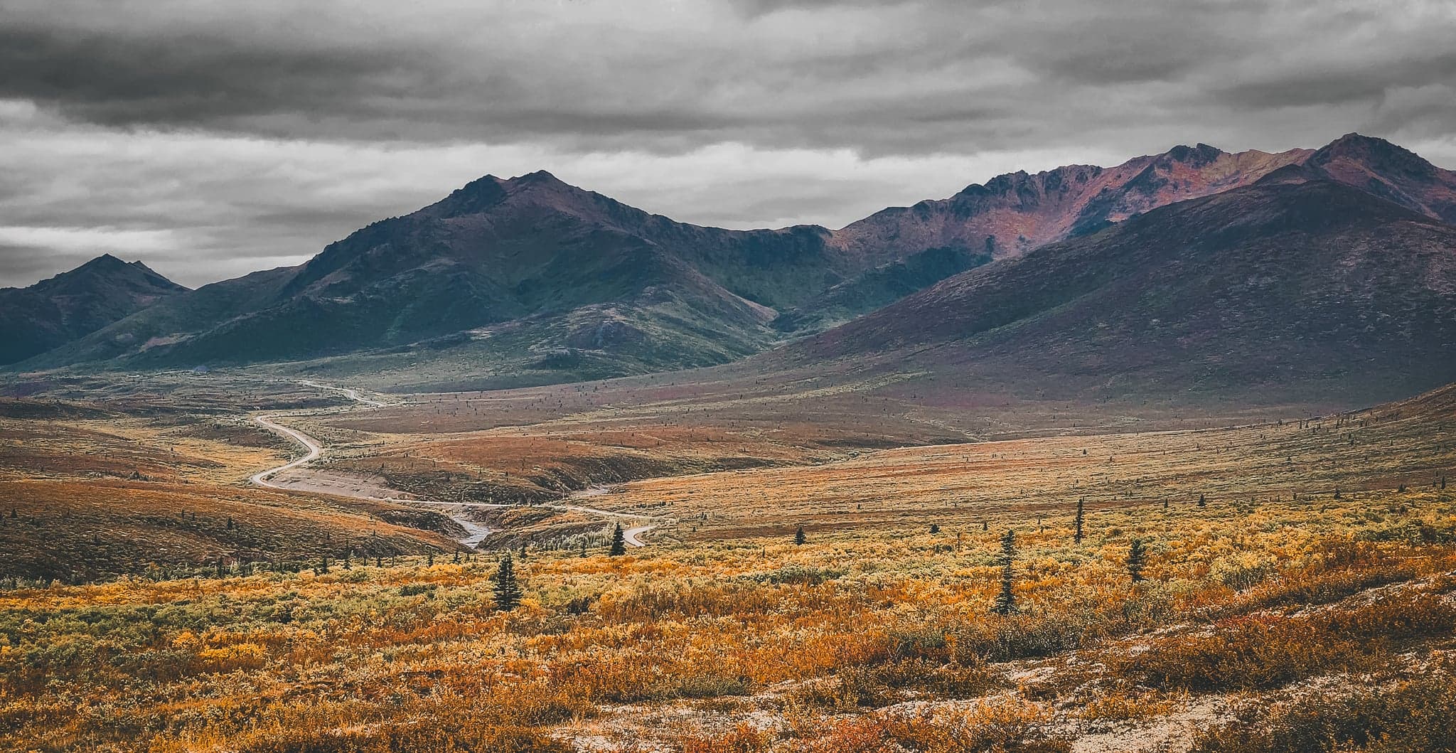 Brooding clouds hover over expansive, golden-hued plains, as a meandering path leads towards rugged mountains cloaked in shadow.