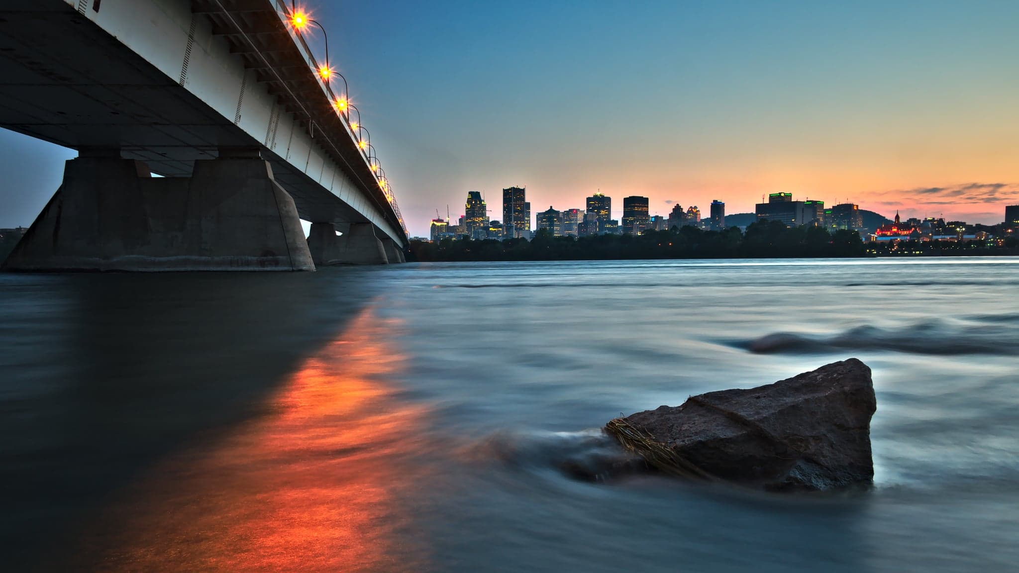 The city skyline emerges against a twilight sky, while bridge lights cast a warm glow over the serene, flowing river.