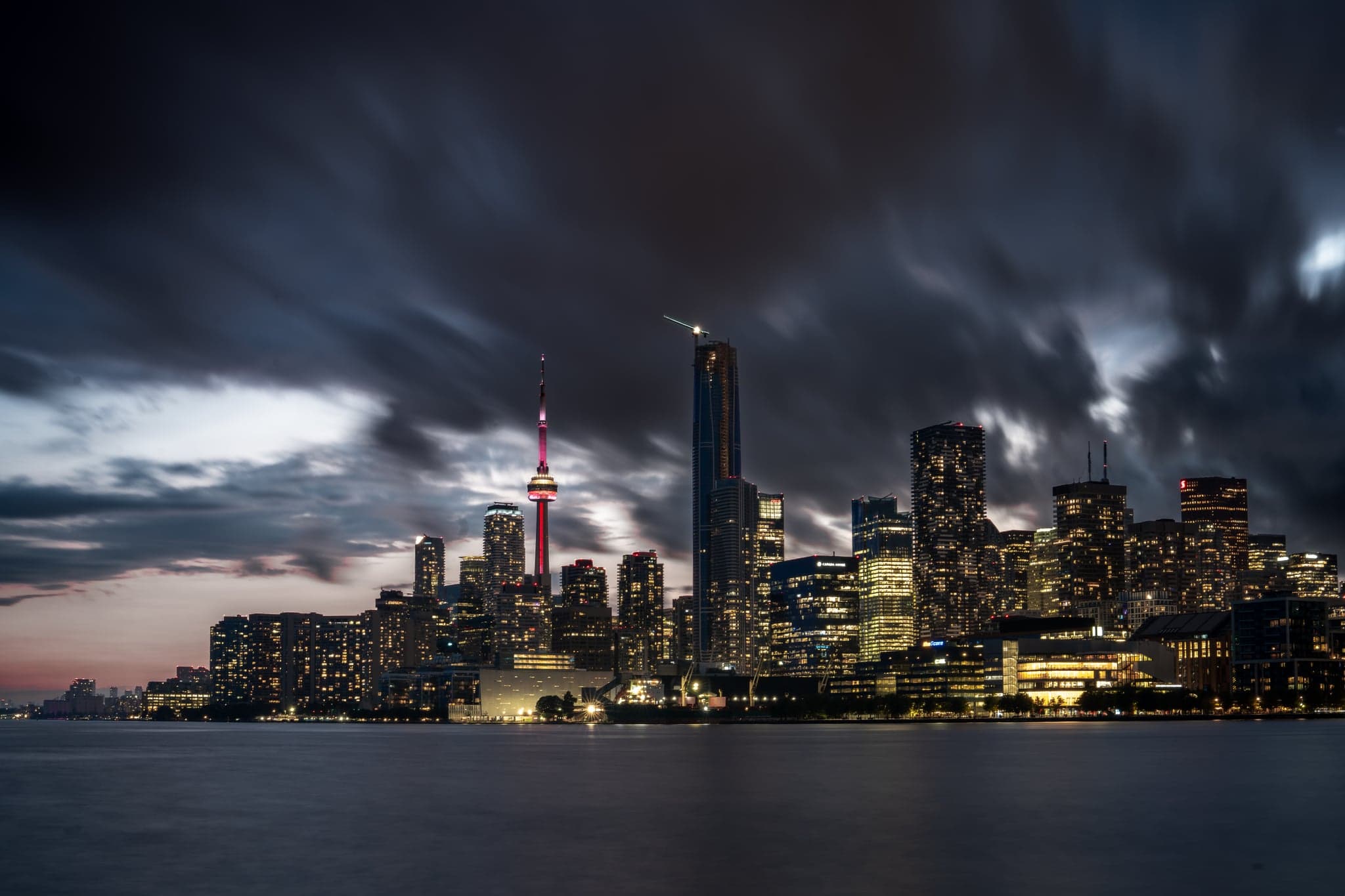 The city skyline glimmers under a moody sky, where streaks of cloud dance above the luminous towers reflecting on calm waters.